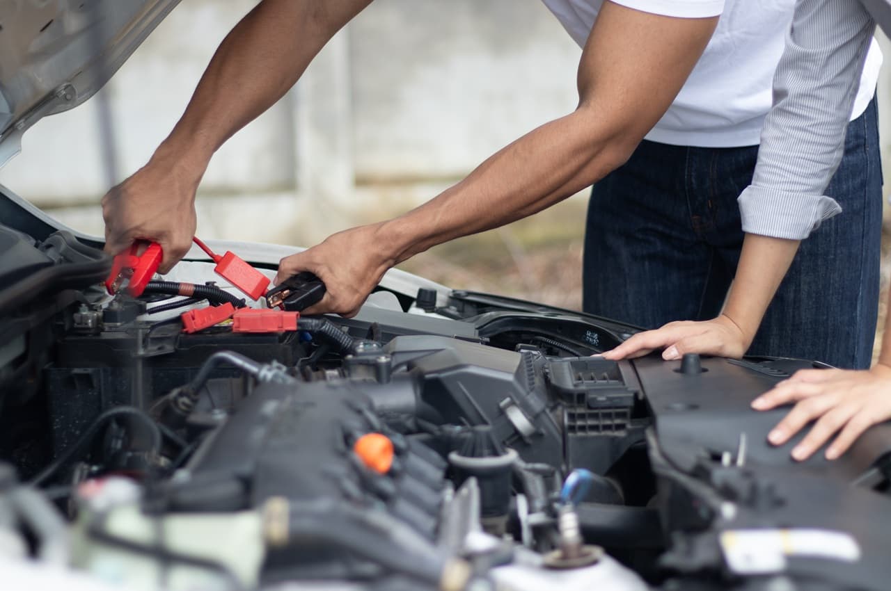 Mãos conectando cabos de bateria no motor de um carro com o capô aberto.