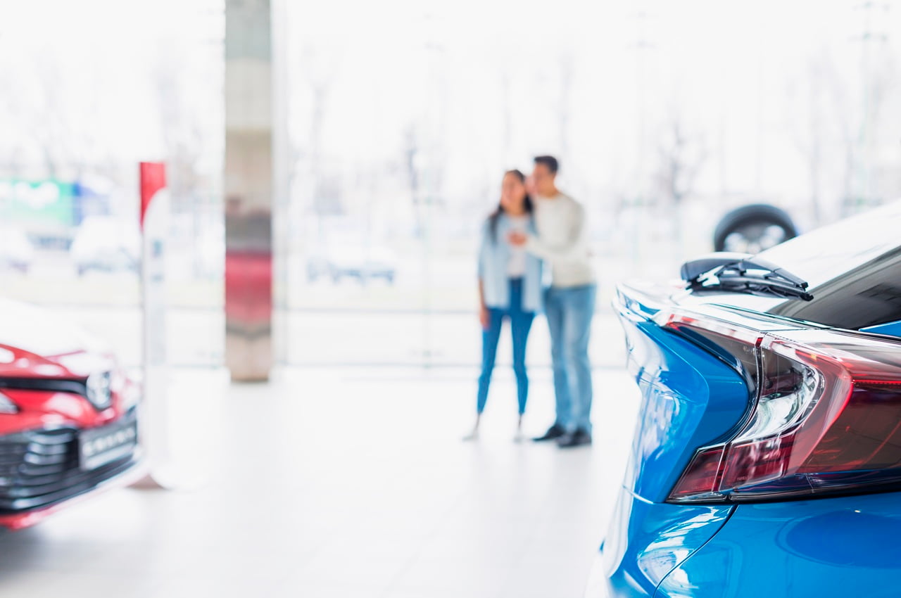 Close na lanterna traseira de um carro azul em uma concessionária, com um casal desfocado em pé ao fundo, observando os veículos.
