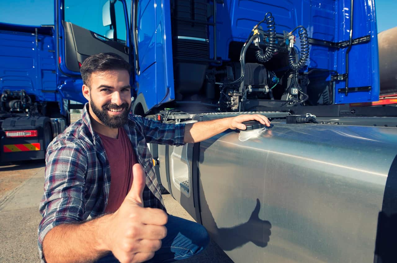 Homem sorridente de camisa xadrez agachado ao lado de um caminhão azul, enquanto faz um sinal de joia com uma das mãos.