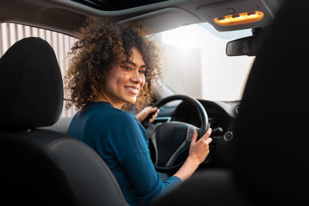 Mulher com cabelo crespo e roupa azul, no banco do motorista de um carro moderno com direção elétrica.