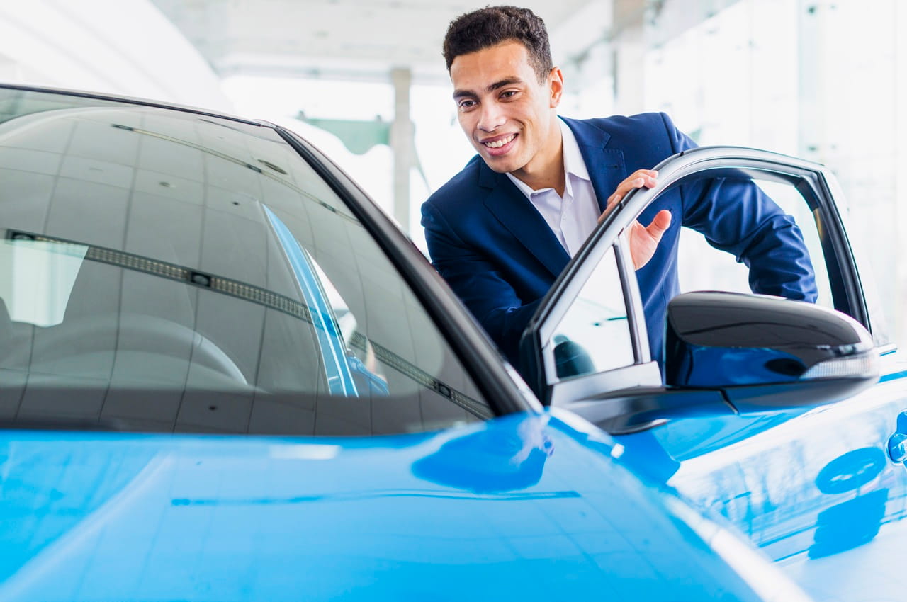 Jovem sorridente em uma concessionária, ao lado de um carro azul novo, comprado via Feirão BV.
