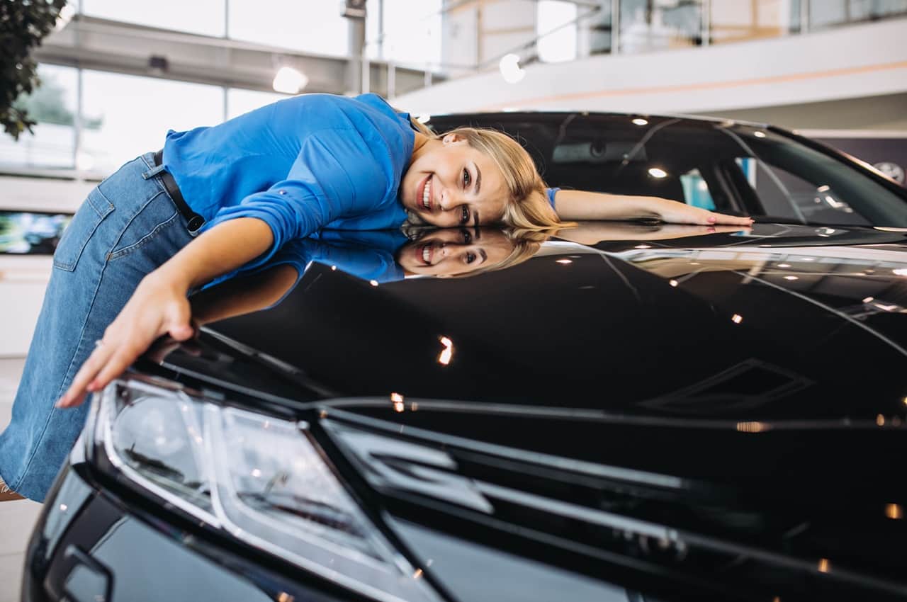 Mulher loira sorridente abraçando o capô de um carro preto brilhante em uma concessionária.
