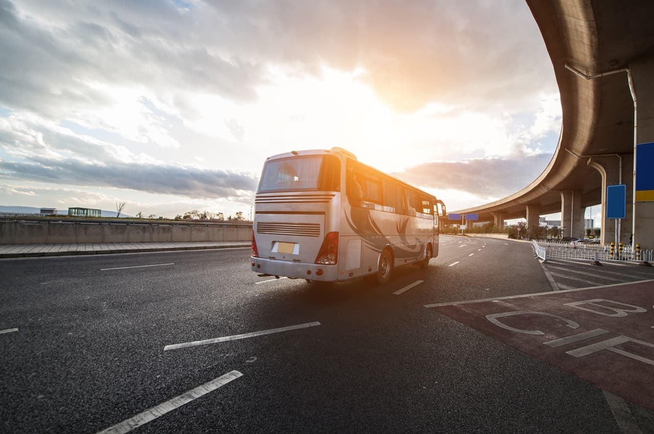 Ônibus branco em movimento na estrada ao pôr-do-sol, com céu parcialmente nublado e cenário urbano ao fundo.