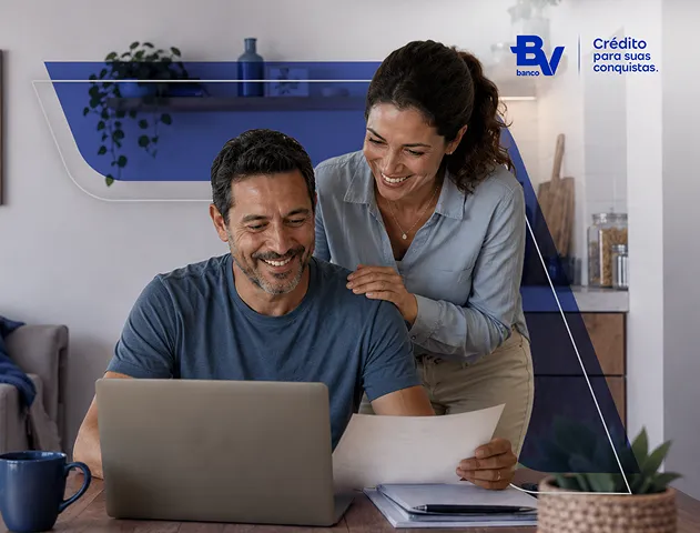 Casal sorrindo. O homem está sentado, trabalhando em seu notebook, e a mulher está em pé ao lado dele, tocando o seu ombro. 