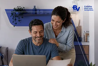 Casal sorrindo. O homem está sentado, trabalhando em seu notebook, e a mulher está em pé ao lado dele, tocando o seu ombro. 