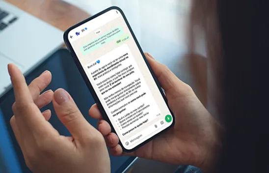 Mão segurando um celular. Na tela do celular, temos uma conversa no WhatsApp.