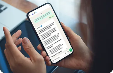 Mão segurando um celular. Na tela do celular, temos uma conversa no WhatsApp.