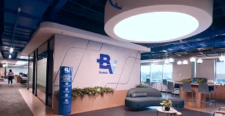 Sala corporativa com sofá, mesas, cadeiras e plantas. Na parede, vemos a logo do banco BV e, ao fundo, uma grande janela. 