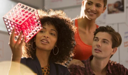 Imagem de uma mulher negra segurando um cubo rosa na mão. Um homem branco e uma mulher branca estão ao lado olhando a mulher negra segurando o cubo rosa.