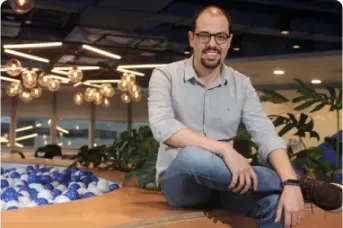 Imagem de um homem branco sentado em uma estrutura de madeira, dentro de um escritório. Ele está sorrindo, usa óculos e está de camisa cinza e calça jeans. Ao lado dele, há uma piscina de bolinhas brancas e azuis, cores que representam a marca do banco BV.