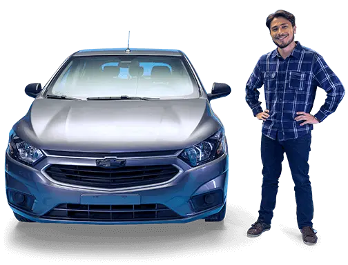 À direita, um homem branco, de cabelos lisos e pretos e barba curta, está em pé apoiando as duas mãos em sua cintura e sorrindo.
Ele está com uma camisa de manga longa xadrez, nas cores azul marinho e branca, calça jeans escura e sapatênis preto.
Do lado direito dele está a frente um carro novo, na cor prata.