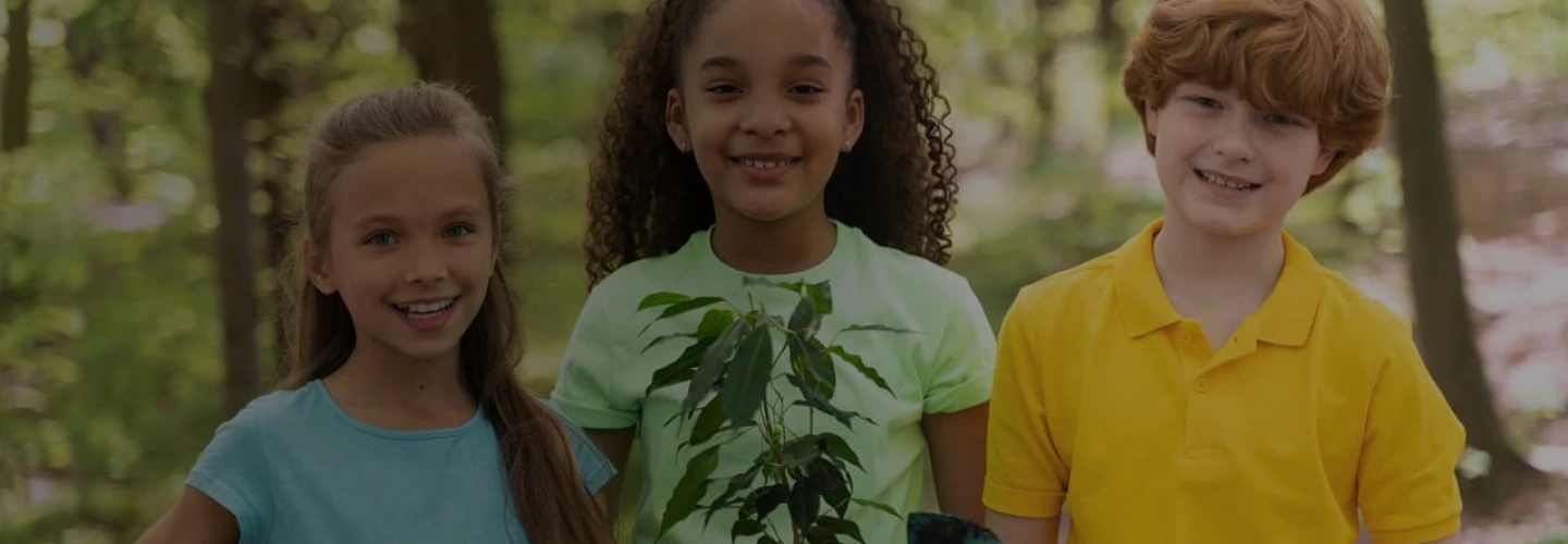 Três crianças sorrindo enquanto uma segura uma planta, com árvores ao fundo.