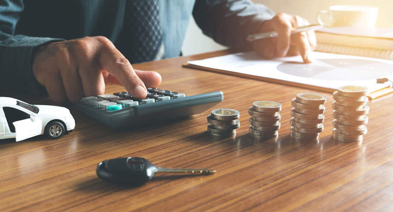 Mãos de um homem fazendo cálculos em uma calculadora. Sobre a sua mesa, há moedas, uma miniatura de um carro e uma chave.