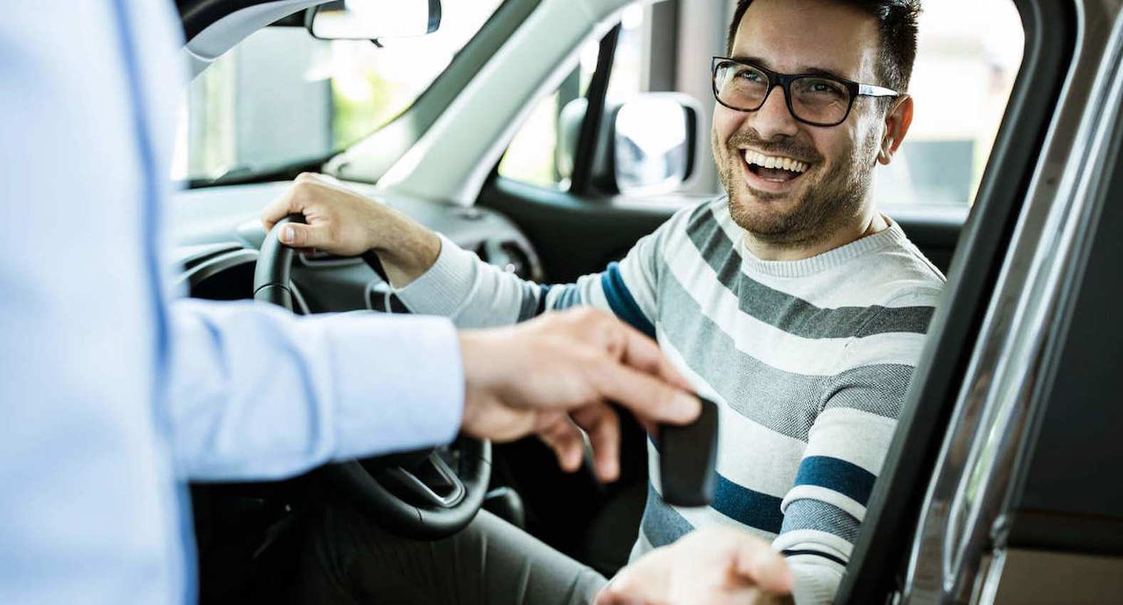Rapaz dentro de um carro em uma concessionária, enquanto financia um carro sem entrada. Ele está no banco do motorista e sorridente.