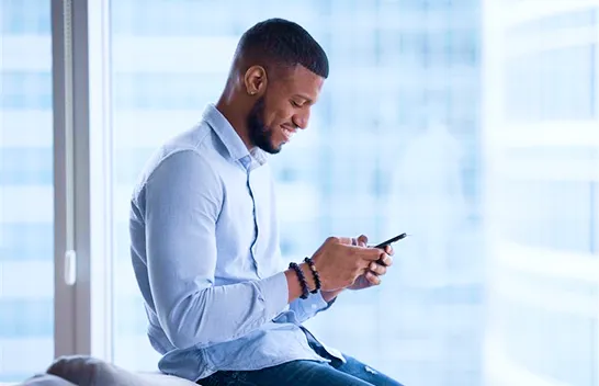 Homem sorridente em ambiente corporativo segurando o celular com as duas mãos.