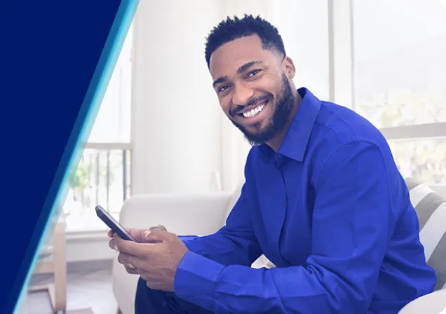 Homem sorridente, com camisa azul, segurando um celular. Ele está sentado em um ambiente iluminado.