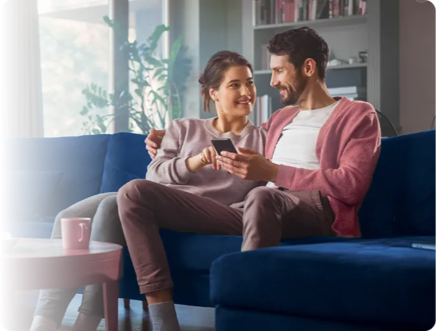 Um casal formado por um homem e uma mulher sentados em um sofá se olhando e mexendo em um celular.
