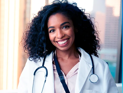 A imagem tem um fundo de prédios desfocados. Em primeiro plano, temos uma mulher jovem, negra, de cabelo ondulado escuro. Ela está sorrindo e veste um jaleco branco e abaixo do jaleco é possível ver uma camisa rosa. Ela está com um estetoscópio em volta do pescoço.