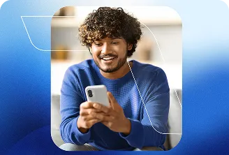 Homem com blusa azul de mangas longas, segura o celular com as duas mãos, olha o aparelho e sorri.