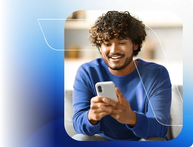 Homem com blusa azul de mangas longas, segura o celular com as duas mãos, olha o aparelho e sorri.