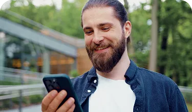 Um homem branco, de barba, usa camisa azul, olha para o celular e sorri.