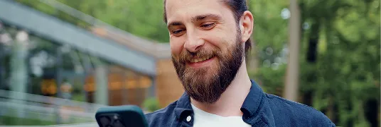 Um homem branco, de barba, usa camisa azul, olha para o celular e sorri.