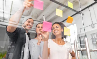 Duas mulheres e um homem em frente a um quadro transparente realizando anotações em post-its.