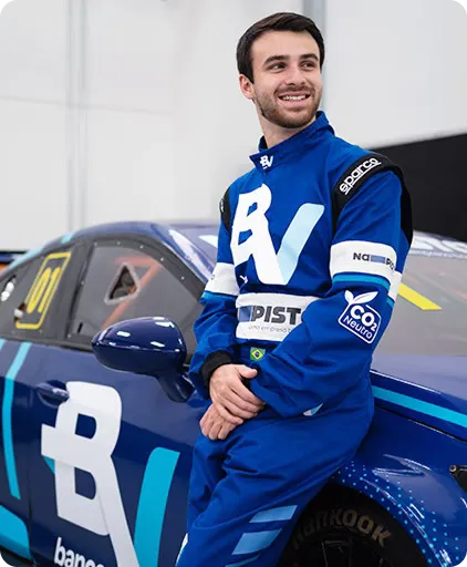 Um homem com roupa de piloto de corrida, encostado em um carro com a logo do banco BV.