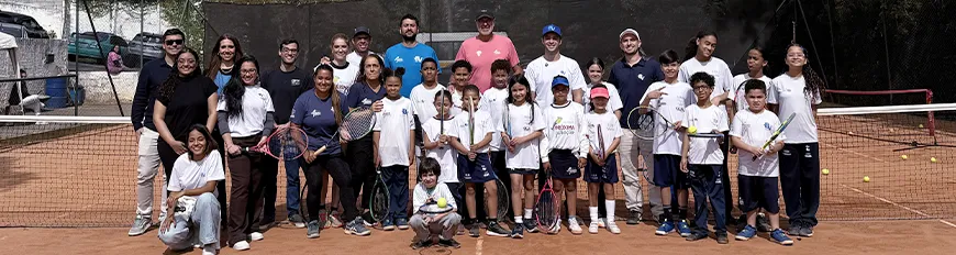 Várias pessoas juntas, adultos, crianças e jovens em uma quadra de tênis, segurando raquetes, posicionados para uma foto.