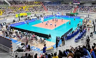 Uma quadra de vôlei, com atletas jogando, e várias pessoas na arquibancada torcendo e assistindo.