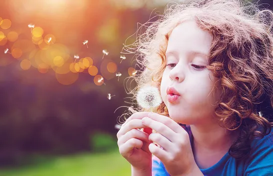 Criança de cabelos encaracolados soprando um dente-de-leão, sementes voando com luz dourada ao fundo.