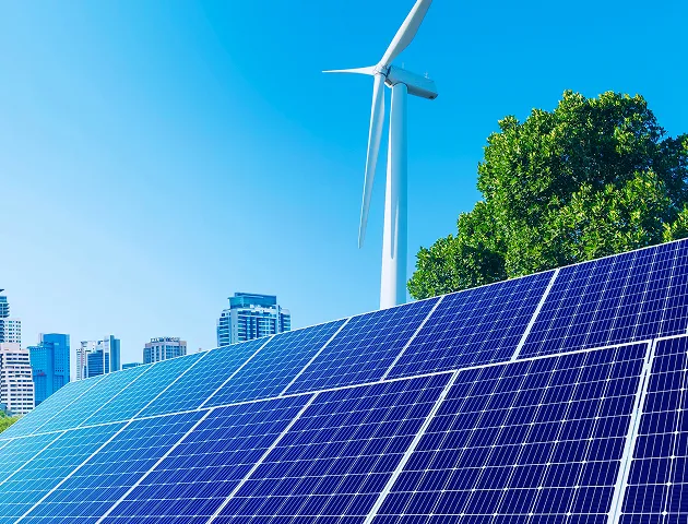 Painel solar em primeiro plano, turbina eólica ao fundo e árvores com prédios sob céu azul.