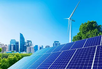 Painel solar em primeiro plano, turbina eólica ao fundo e árvores com prédios sob céu azul.