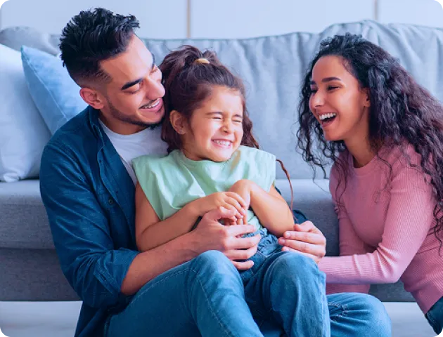 Fotografia de homem e mulher sentados no chão da sala. O homem segura uma menina no colo, e os três sorriem juntos.