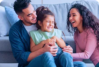 Fotografia de homem e mulher sentados no chão da sala. O homem segura uma menina no colo, e os três sorriem juntos.