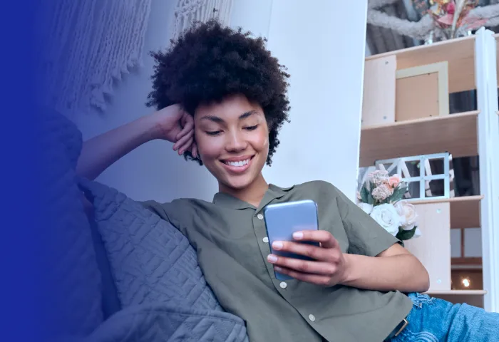 Mulher sorrindo recostada no sofá, usando camisa verde, segurando um celular em ambiente interno com estante ao fundo.