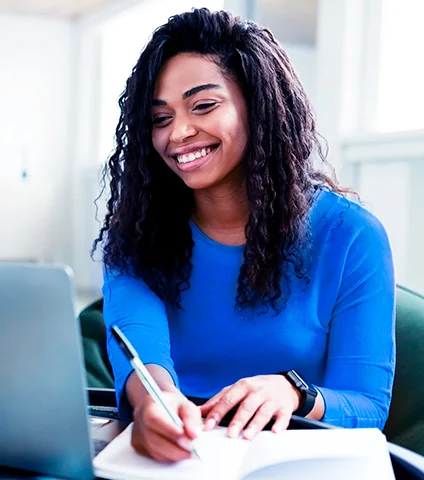 A imagem mostra uma garota negra de cabelos longos, que está sentada em uma poltrona verde, sorrindo, em frente a um notebook. Ela veste uma blusa de manga longa na cor azul. Com a mão esquerda ela segura uma caneta preta, enquanto escreve nos papéis que estão apoiados na mesa, e é possível ver um relógio preto no pulso direito. O fundo da imagem é branco com janelas.