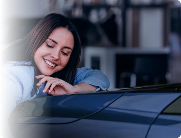 Mulher de olhos fechados sorrindo e apoiada no capô de um carro azul escuro.