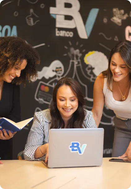 Três mulheres sorrindo e olhando para um laptop. Ao fundo, vemos uma parede preta com grafismos e a logo do BV.