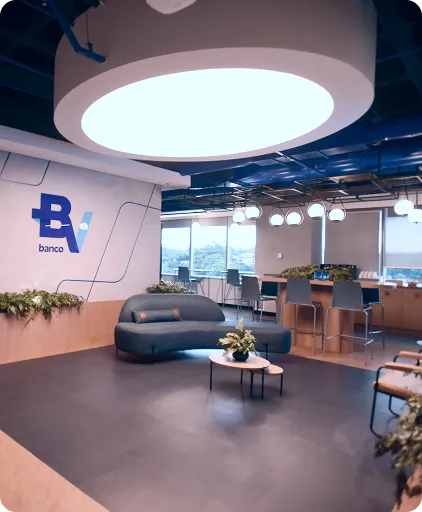 Sala corporativa com sofá, mesas, cadeiras e plantas. Na parede, vemos a logo do banco BV e, ao fundo, uma grande janela. 