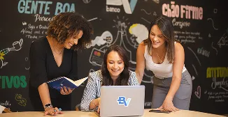 Três mulheres sorrindo e olhando para um laptop. Ao fundo, vemos uma parede preta com grafismos e a logo do BV.