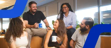 Um homem e duas mulheres sentados, sorrindo de maneira descontraída e segurando uma caneca com a logo do banco BV. 