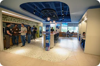 Copa do escritório do banco BV, onde apresenta um balcão central grande com algumas banquetas com o logo do banco logo acima, algumas mesas separadas a direita, e uma pia grande onde várias pessoas estão interagindo.