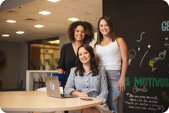 Em um fundo de ambiente corporativo, uma mulher sentada está sorrindo e em sua frente está uma mesa com um notebook, atrás dela estão duas mulheres também sorrindo.