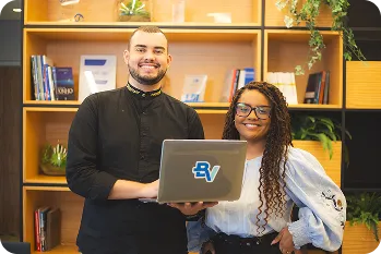 Um homem sorrindo e segurando um notebook ao lado de uma mulher sorrindo, ao fundo uma estante com livros e plantas.