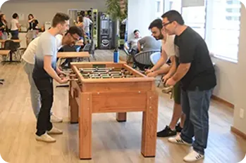 Quatro homens estão em pé ao redor de uma mesa de pebolim jogando, ao fundo da imagem, outras pessoas conversam.