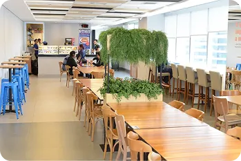 Área de convivência com mesas de madeira, cadeiras, bancos azuis e plantas. Ao fundo, pessoas estão sentadas comendo.