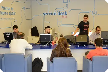 Três pessoas sentadas de costas e três, em frente a uma bancada, trabalhando. Ao fundo, uma pessoa de camiseta preta em pé.