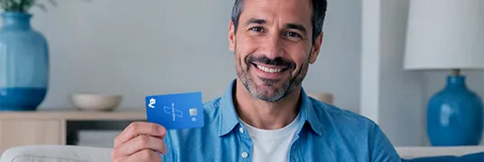 Homem branco, vestindo uma camisa azul, sentado em um sofá. Ele sorri enquanto segura um cartão em uma das mãos.