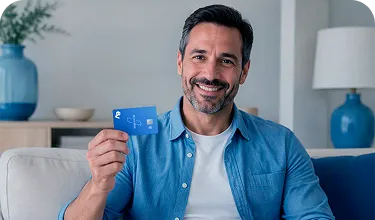Homem branco, vestindo uma camisa azul, sentado em um sofá. Ele sorri enquanto segura um cartão em uma das mãos.